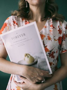 Portrait of a woman holding a booklet in a floral dress, set against a dark background.
